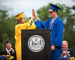 A blue-robed graduate gives a high-five to a yellow-robed graduate at a podium with a circular seal during an outdoor graduation ceremony.