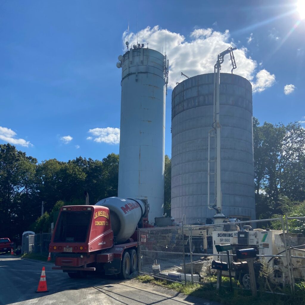 Two tall cylindrical silos on a construction site next to a red cement mixer truck under a bright blue sky.