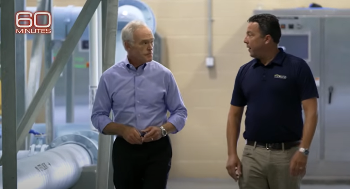 Two men walk through an industrial facility with large pipes during a 60 Minutes interview about cybersecurity threats.