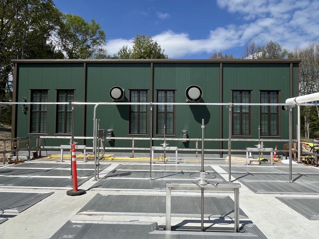 Green industrial building with circular vents, exposed pipes, and safety railings in a paved outdoor utility area.