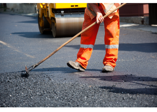 Road construction worker in orange safety gear spreads hot asphalt with a rake, with a roller machine in the background.