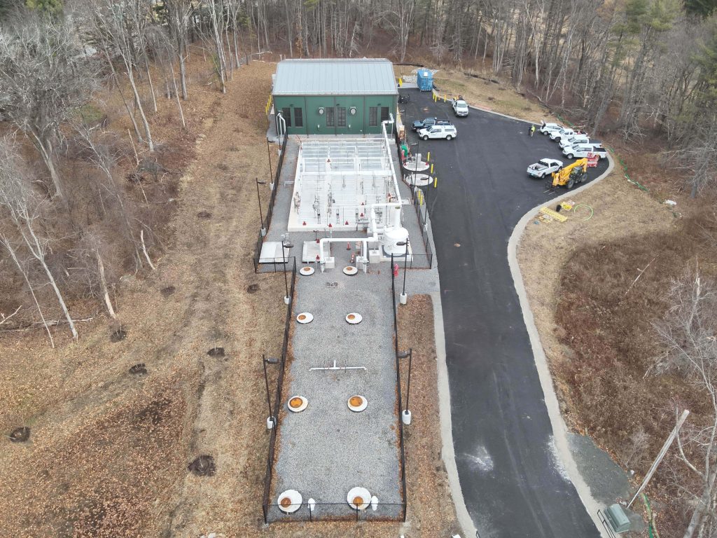 Aerial view of a small sewer facility with a long gray building, a green-roofed auxiliary structure, a paved driveway, and several parked vehicles, all surrounded by trees.