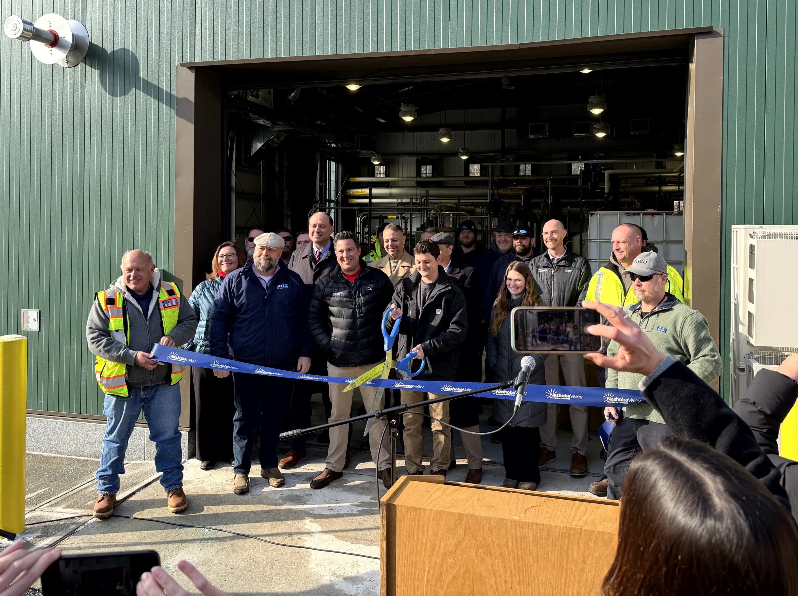 Officials and workers in safety vests stand behind a blue ribbon at the entrance of an industrial facility during a sewer system ribbon-cutting ceremony.